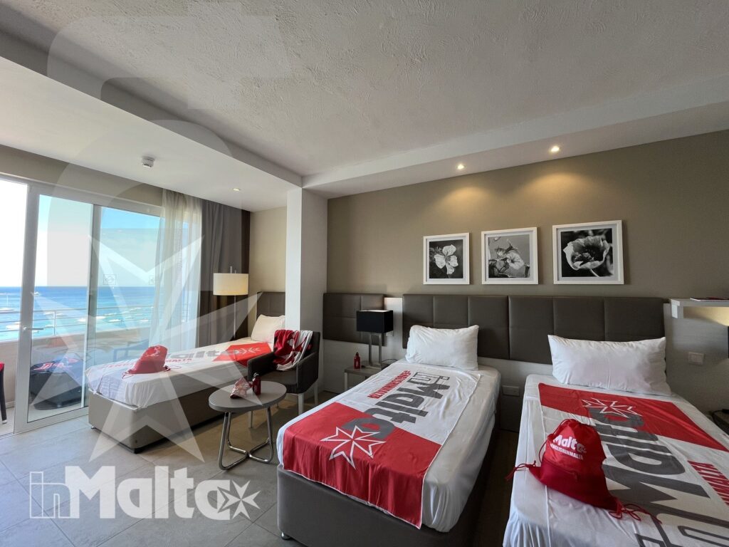 Camera luminosa con due letti singoli, divano, balcone e vista sul mare, pensata per corsisti durante una vacanza studio a Malta.