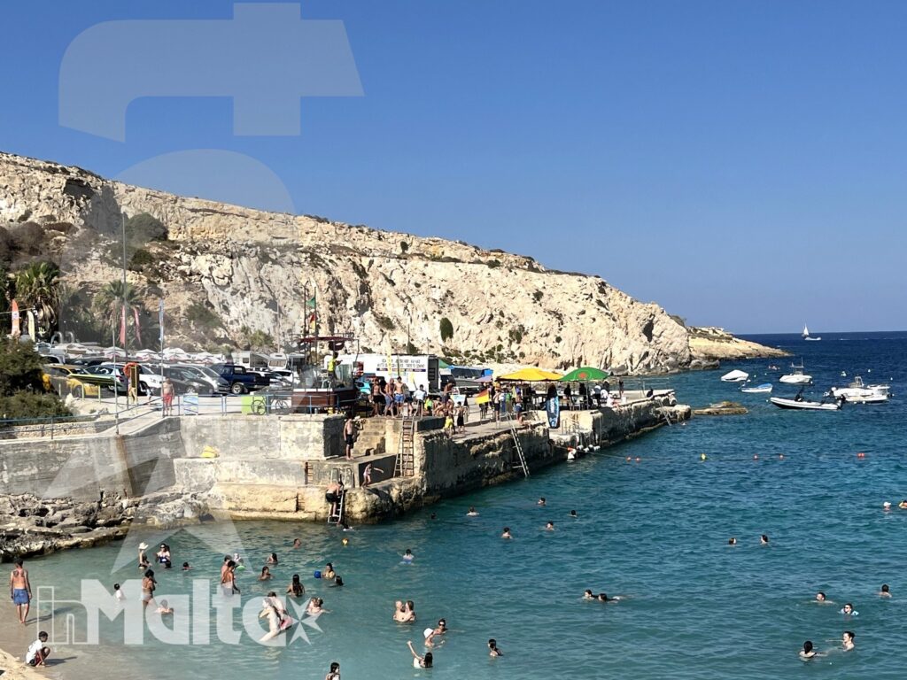 Baia di Hondoq Bay tra barche, pontile e scogliere Baia maltese animata da bagnanti, pontile e barche ormeggiate, incorniciata da una luminosa scogliera calcarea.