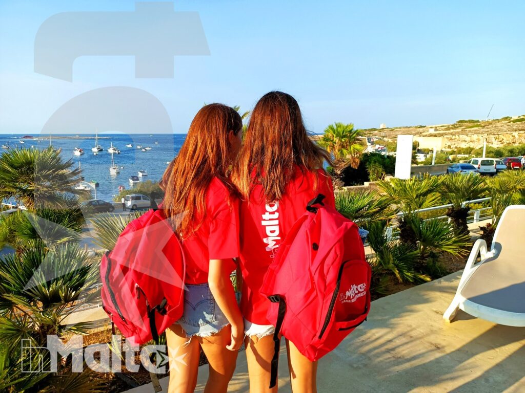 Due studentesse con zaini rossi osservano una baia mediterranea, con vegetazione e barche sul mare durante il soggiorno a Malta.