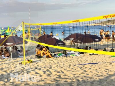 Campo da beach volley sulla sabbia con giocatori, pubblico, ombrelloni e lettini, nel vivace ambiente di una località balneare.