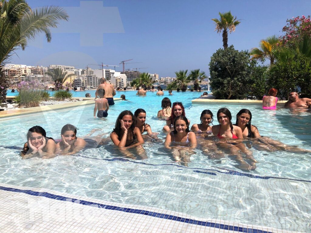 Gruppo di studenti durante il soggiorno a Malta Gruppo numeroso di studenti sorride immerso nella piscina, con il resort e la costa mediterranea sullo sfondo.