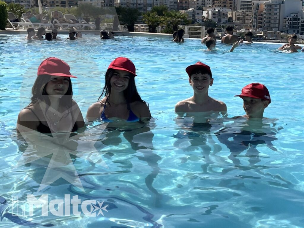 Quattro studenti con cappellini rossi sorridono immersi nella piscina, davanti a uno skyline urbano durante il soggiorno a Malta.