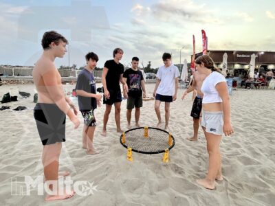 Partecipanti raccolti attorno a una rete da racchettoni sulla spiaggia, tra socializzazione e attività all'aperto organizzate da inMalta.
