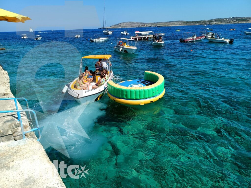 Attività in gommone per studenti nella baia di Gozo Studenti salgono su un gommone accanto a una ciambella gonfiabile verde, nelle acque turchesi della baia di Gozo.