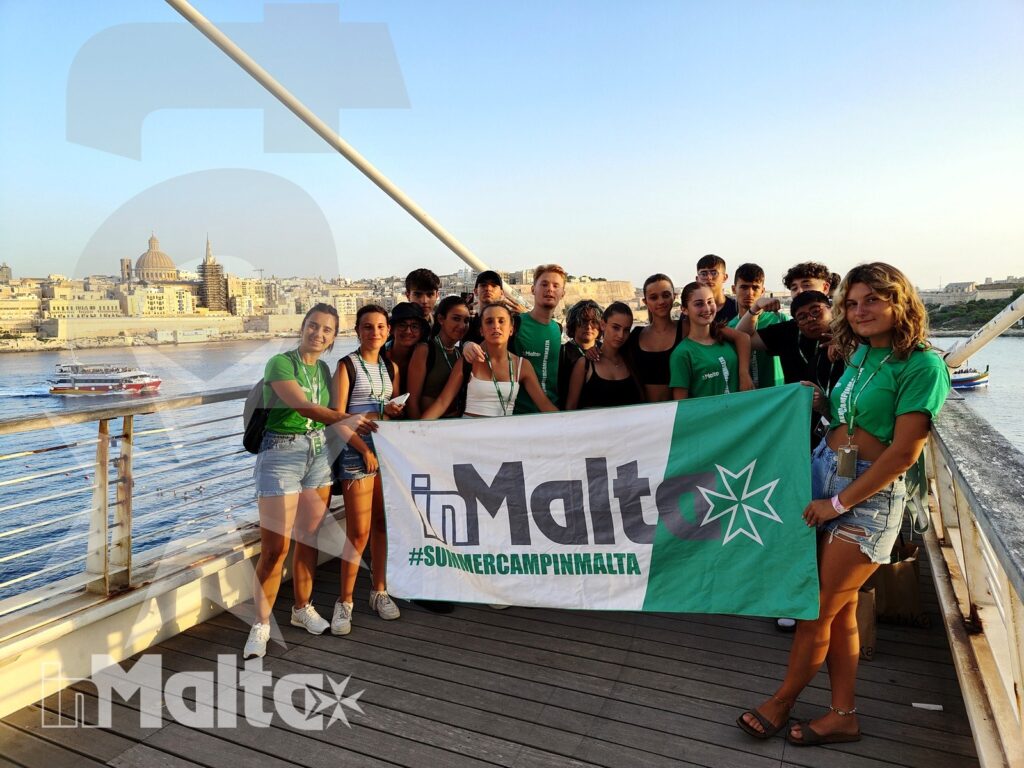 Gruppo di studenti posa sul ponte di un traghetto con una bandiera, mentre sullo sfondo si vedono il porto e il panorama urbano di Malta.