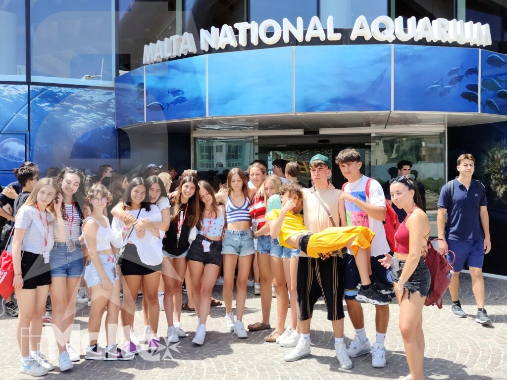 Gruppo di studenti davanti al Malta National Aquarium Gruppo numeroso di studenti sorride davanti all'ingresso del Malta National Aquarium durante un'attività organizzata a Malta.