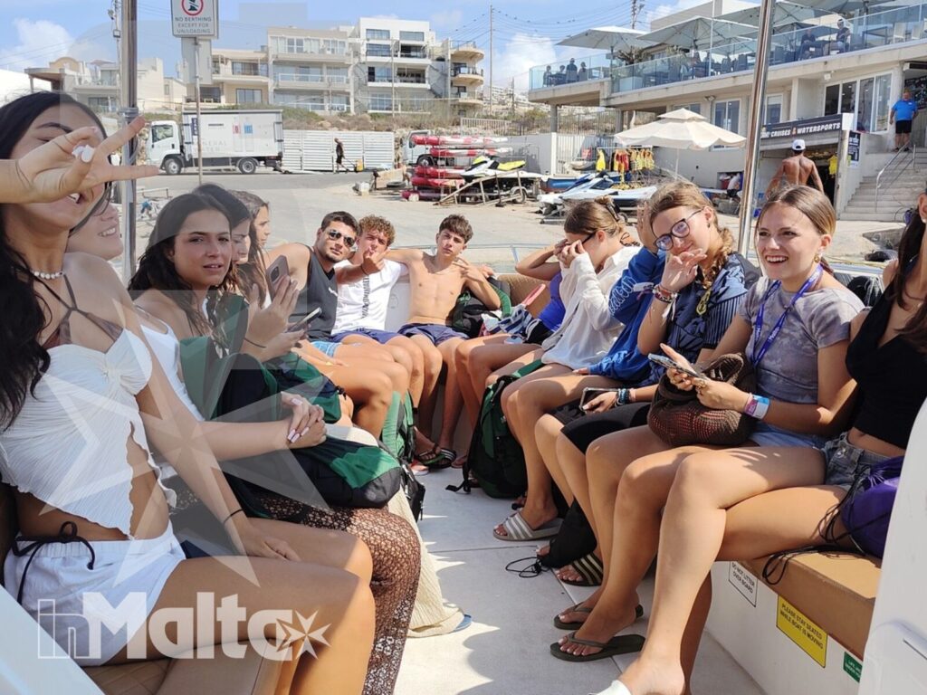 Gruppo internazionale di studenti sorride seduto su una barca durante un'escursione in un porto maltese soleggiato.