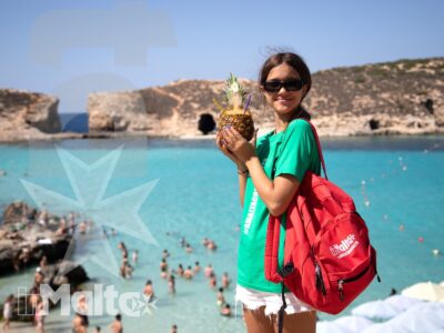 Studentessa sorridente con zaino rosso e ananas in mano posa davanti a una baia turchese affollata durante l'escursione al mare.