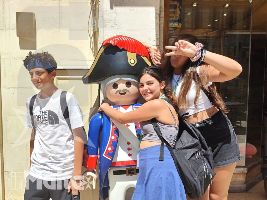 Tre studenti sorridenti posano accanto a una statua colorata davanti a una facciata urbana maltese durante una visita a Malta.