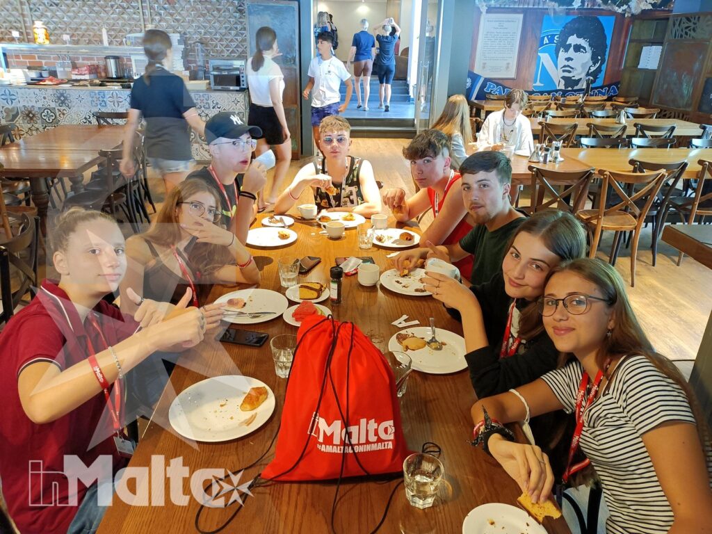 Gruppo numeroso di studenti mangia e conversa attorno a un tavolo apparecchiato, con zaini e cucina a vista sullo sfondo.