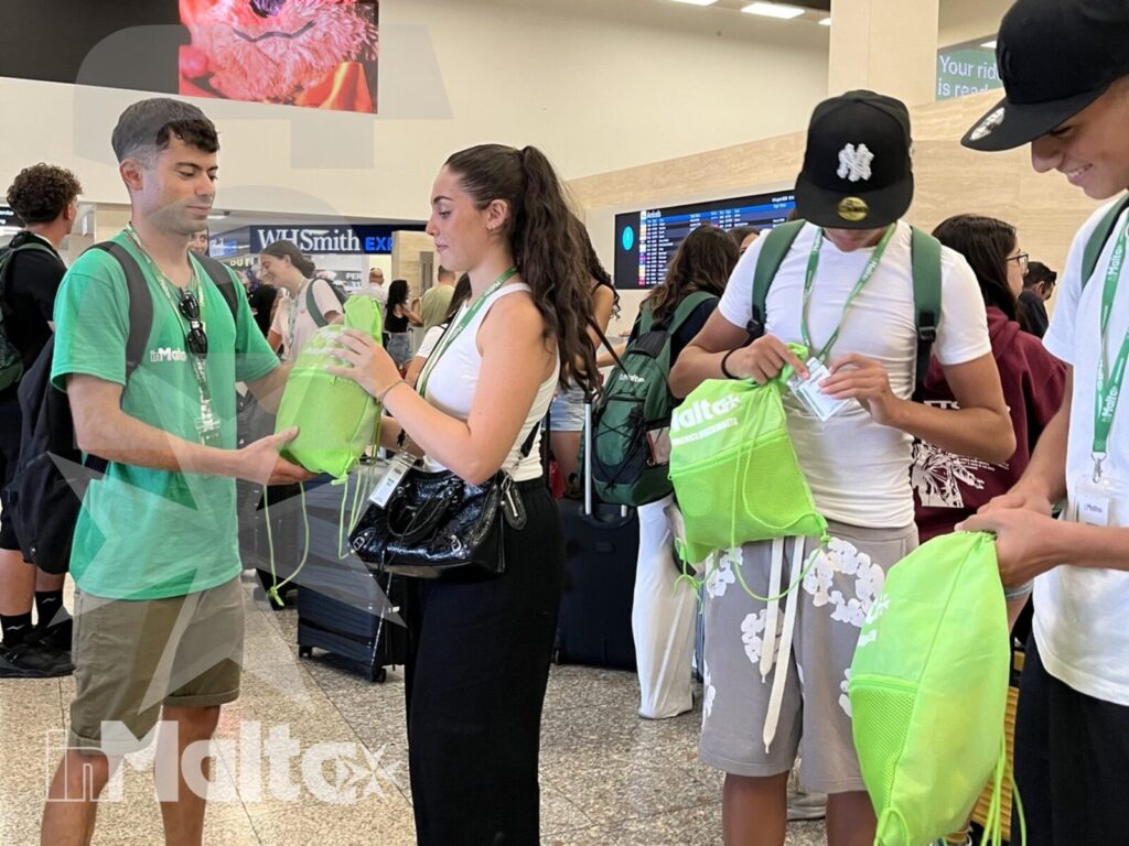 Tutor e studenti durante l'accoglienza per il soggiorno studio Studenti internazionali ricevono borse verdi da un tutor in uno spazio interno affollato durante l'accoglienza inMalta.