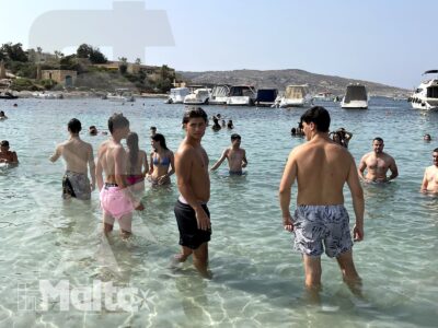 Gruppo numeroso di studenti fa il bagno nelle acque basse e limpide di Santa Maria Beach, tra barche e costa maltese.
