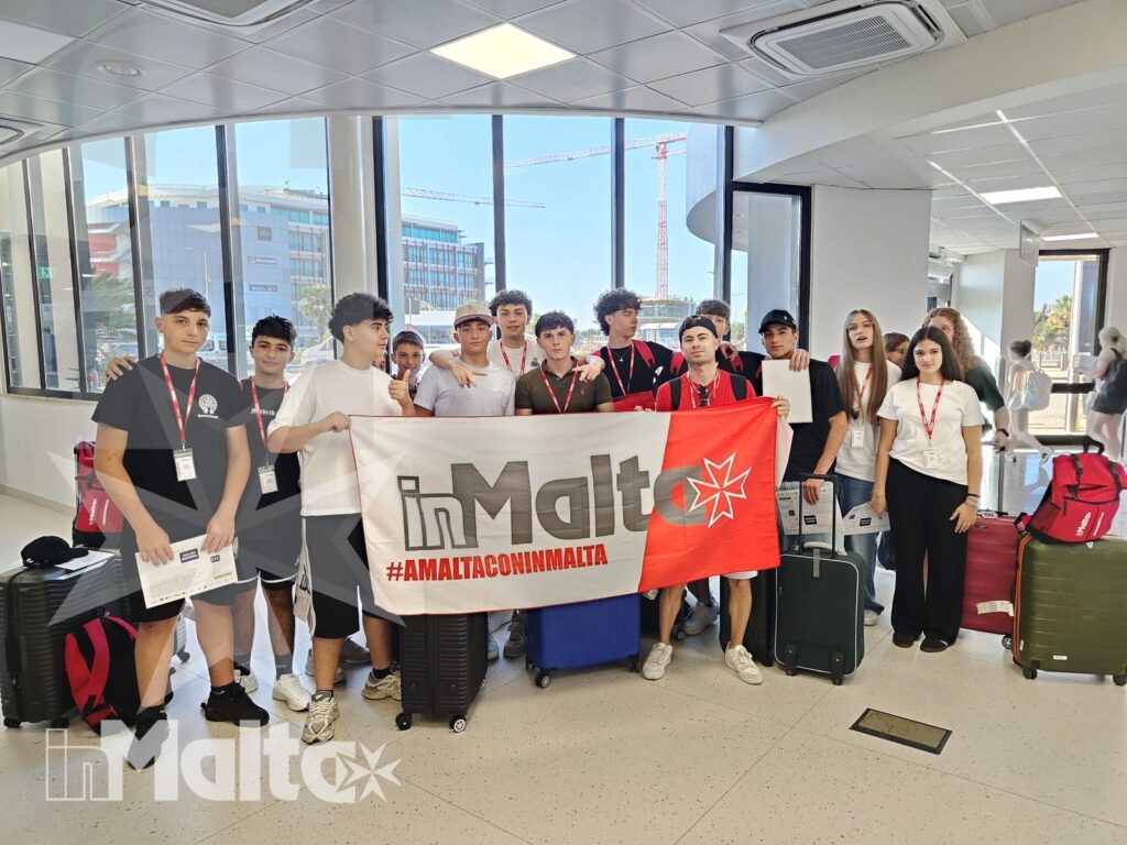 Gruppo di studenti posa in aeroporto con bagagli e striscione, mentre attraverso le vetrate si intravede il paesaggio urbano.