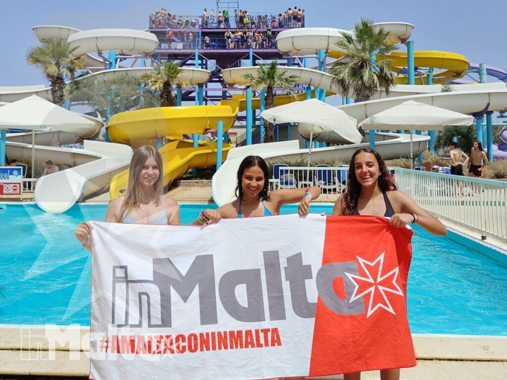 Tre giovani partecipanti mostrano una bandiera inMalta davanti a piscine, palme e scivoli colorati del parco acquatico.