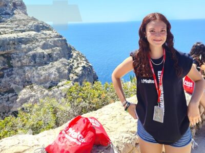 Studentessa con badge e borsa rossa posa sulla scogliera, con il mare blu sullo sfondo durante l'escursione a Malta.