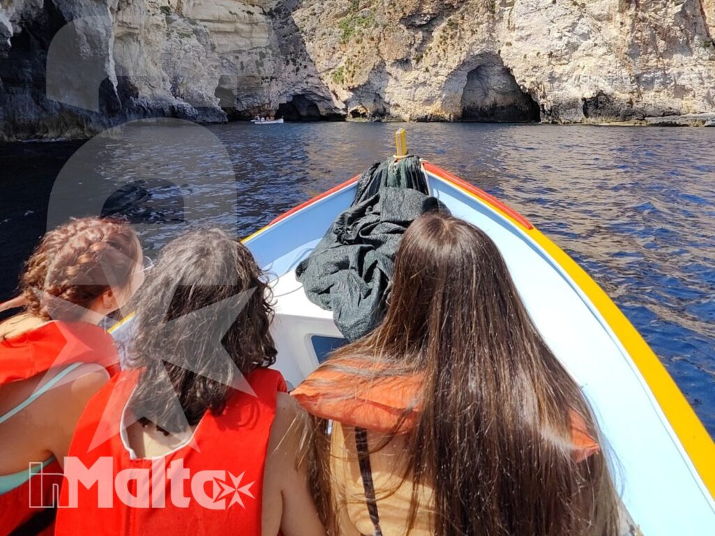 Tre studentesse osservano una grotta marina da una barca colorata, tra scogliere calcaree e acqua blu durante l'escursione alla Blue Grotto.