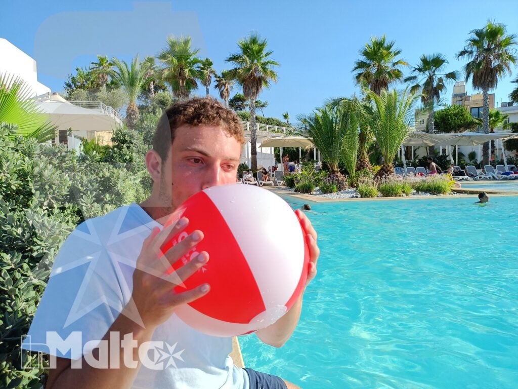 Giovane gioca con un pallone gonfiabile rosso e bianco a bordo piscina, tra palme e vegetazione soleggiata.