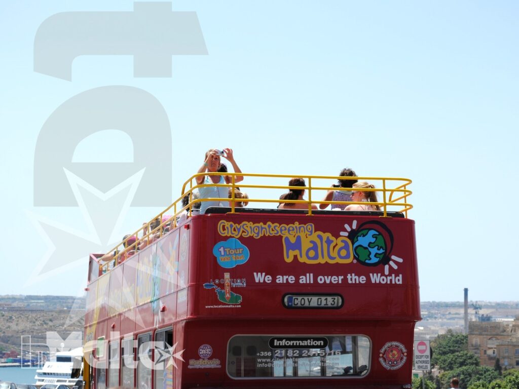 Autobus turistico rosso con passeggeri sul piano scoperto e panorama urbano costiero, durante un'escursione a Malta.