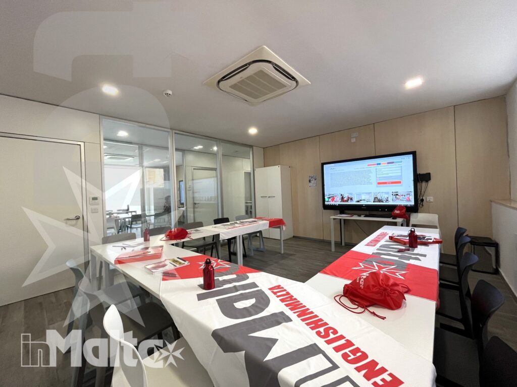 Sala didattica luminosa con tavoli a U, bottiglie, materiali e schermo digitale acceso, predisposta per una lezione di studenti.