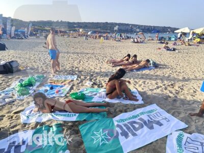 Studenti sdraiati e seduti sulla sabbia con teli promozionali, immersi nel panorama costiero maltese.