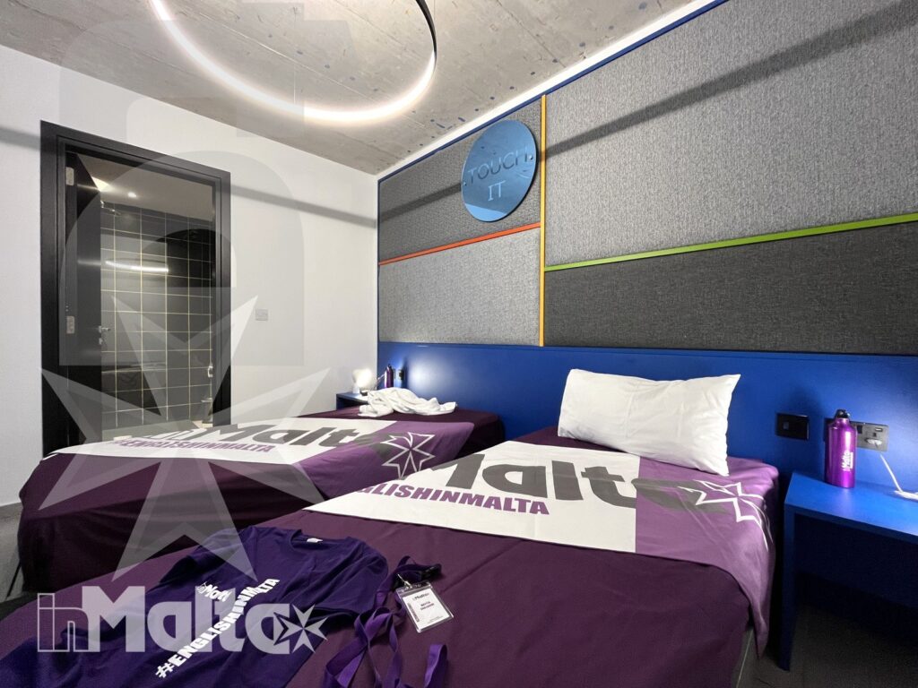 Camera doppia per studenti durante un soggiorno studio a Malta Camera doppia moderna con due letti singoli, copriletti viola e parete decorativa, pronta per accogliere studenti in soggiorno studio a Malta.