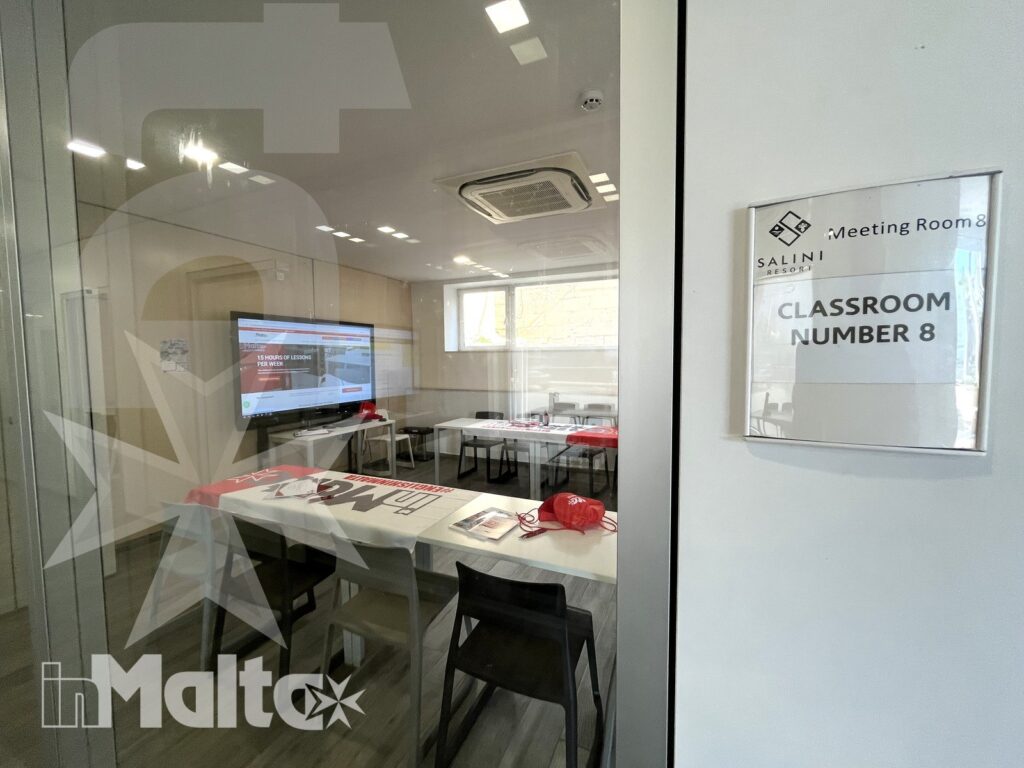 Aula vetrata vuota con monitor acceso, tavoli per lezioni collaborative e cartello Classroom Number 8 visibile all'ingresso.