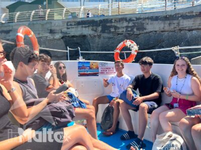 Gruppo internazionale di studenti rilassati siede su una barca da escursione, con salvagenti arancioni e il mare maltese intorno.