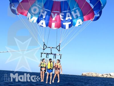 Tre giovani sono sospesi sopra il mare durante il parasailing, sotto una vela colorata con grafica di Stitch e costa rocciosa sullo sfondo.