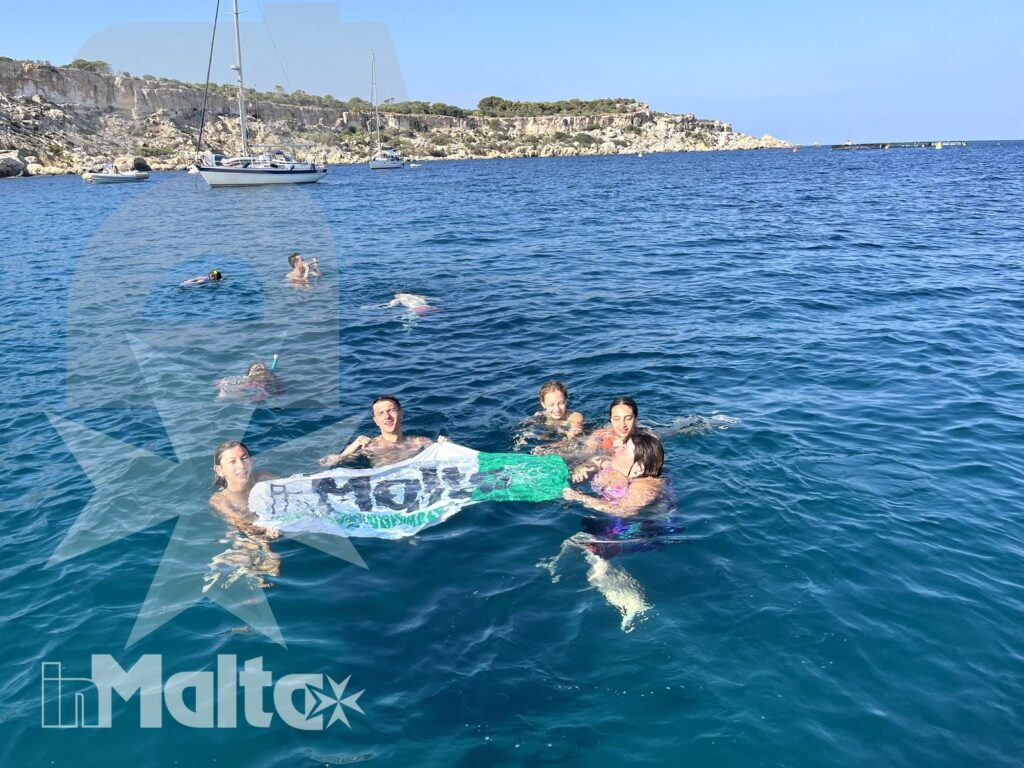 Studenti fanno il bagno insieme in una baia di Malta, raccolti attorno a un materassino tra barche e costa rocciosa.