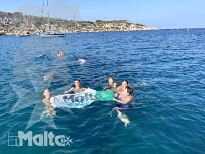 Studenti fanno il bagno insieme in una baia di Malta, raccolti attorno a un materassino tra barche e costa rocciosa.