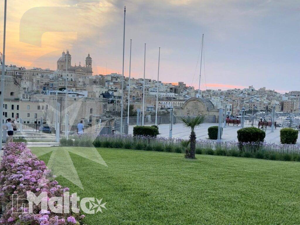 Giardino fiorito con vista sul porto di Malta Giardino fiorito con prato curato, porto e città storica sullo sfondo al tramonto, panorama suggestivo di un viaggio a Malta.