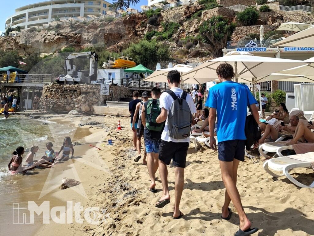Gruppo di studenti cammina sulla sabbia verso il mare, davanti a una struttura sulla scogliera di Golden Bay.
