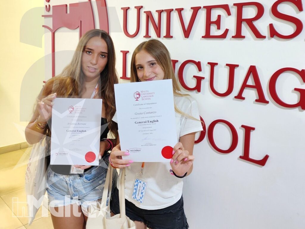 Studentesse mostrano gli attestati al termine del corso a Malta Due studentesse sorridono mostrando i certificati davanti alla parete identificativa della Malta University Language School, dopo il corso di lingua.