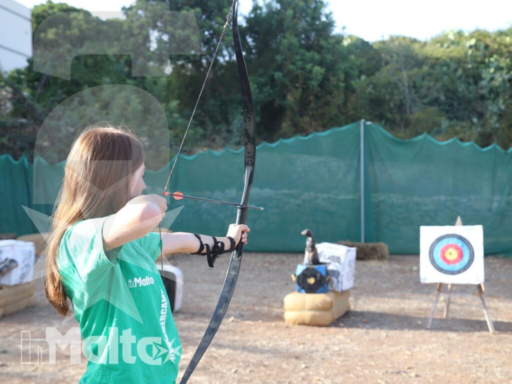 Studentessa con maglietta verde tende l'arco verso un bersaglio durante un'attività all'aperto a Malta.