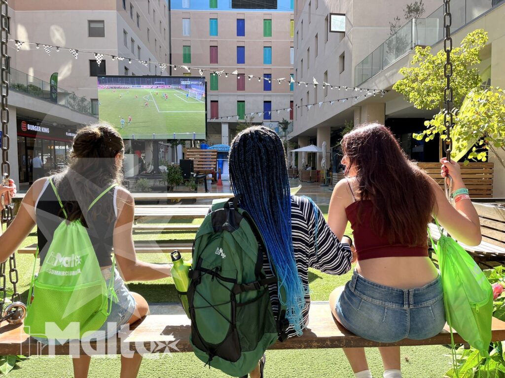 Tre studentesse osservano una partita su un grande schermo dall'altalena di un'area comune all'aperto.