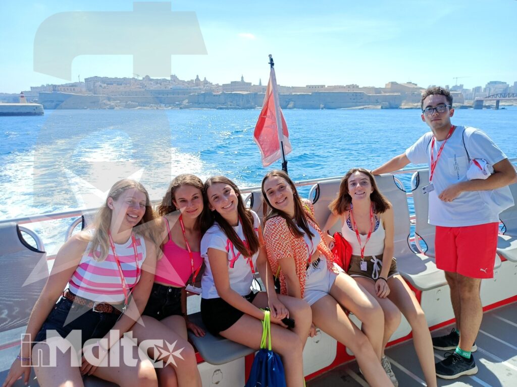 Sei studenti sorridono a bordo di una barca, con il porto e la costa fortificata di Malta sullo sfondo dell'escursione.