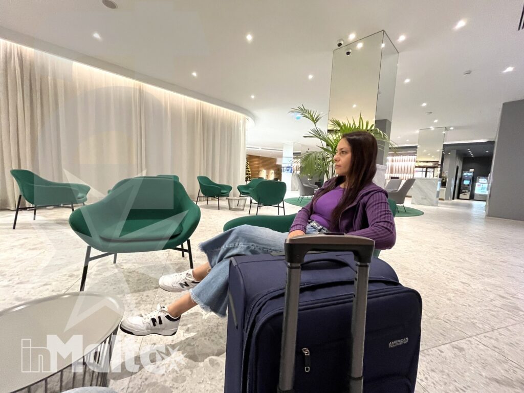 Viaggiatrice con valigia nella lounge dell'alloggio a Malta Giovane viaggiatrice attende seduta in una lounge moderna con la valigia accanto, durante una pausa negli spazi comuni della struttura.