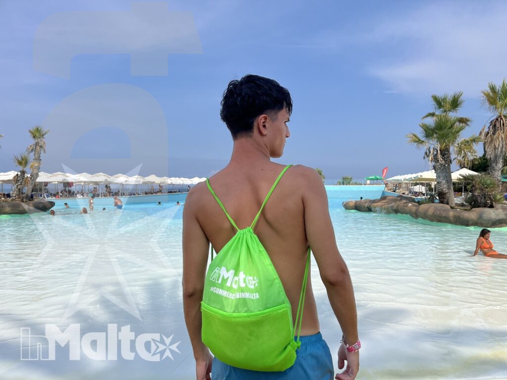 Un giovane con zaino verde cammina verso una laguna balneabile tra palme, ombrelloni e altri ospiti del resort.