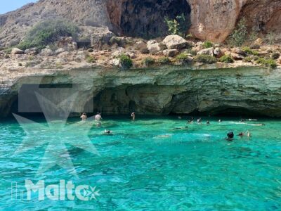 Acqua turchese e trasparente davanti a una grotta marina maltese, con nuotatori tra le rocce durante un'escursione in mare.