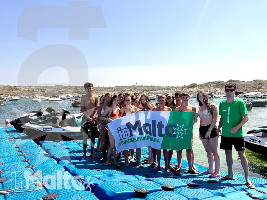 Studenti riuniti su un pontile galleggiante mostrano l'insegna della scuola, mentre alcune moto d'acqua attendono nella baia.