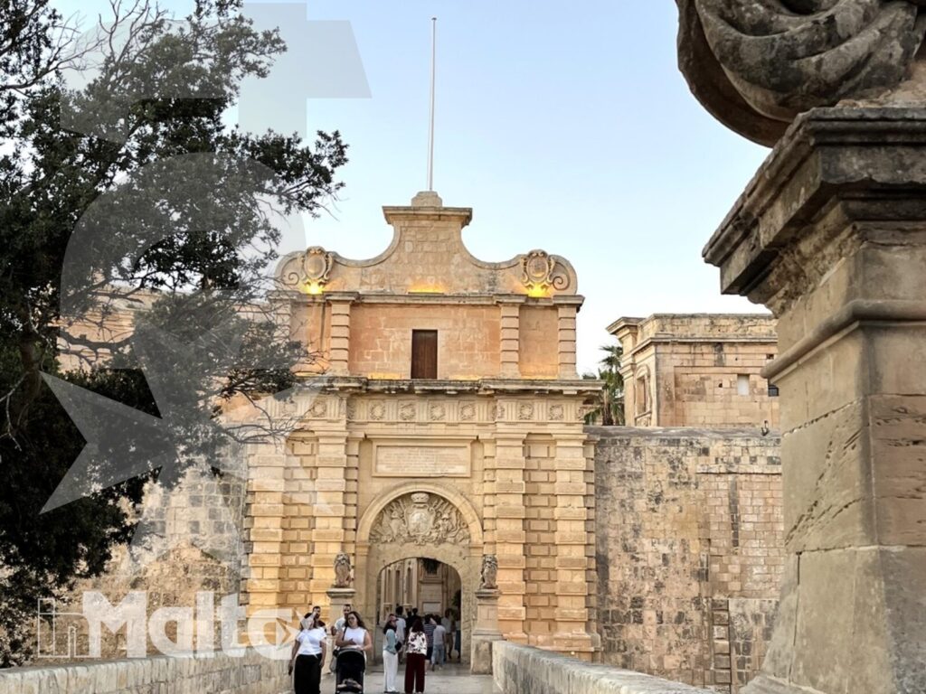 Partecipanti in visita alla porta storica di Mdina Visitatori attraversano un ponte in pietra verso una porta storica di Mdina, tra sculture e architettura maltese.