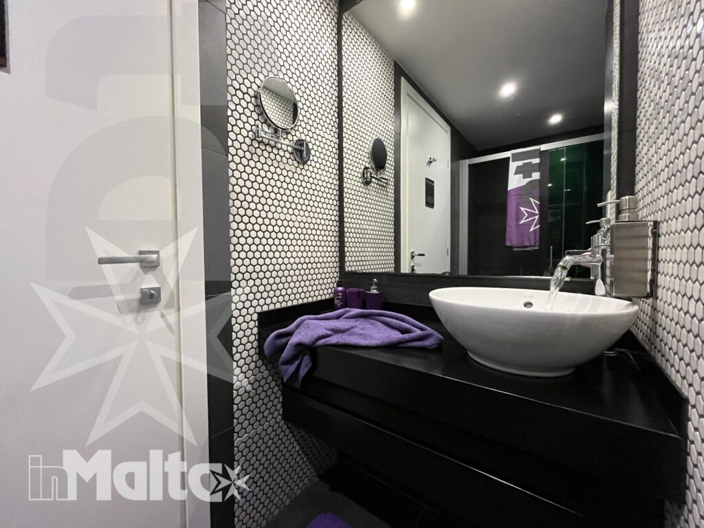 Bagno moderno dell'alloggio con dettagli viola per gli ospiti Bagno moderno con lavabo a bacinella, piastrelle esagonali, mobile scuro, specchi circolari e asciugamani viola coordinati.