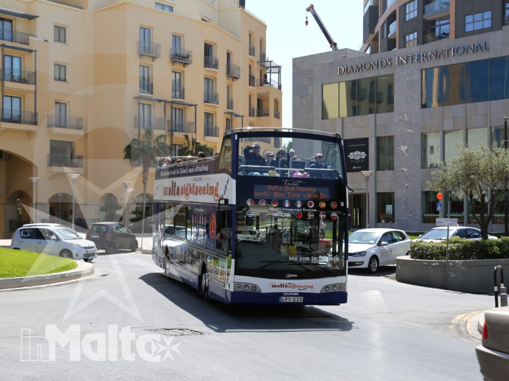 Autobus turistico a due piani attraversa una strada urbana con edifici colorati, nel contesto di un soggiorno a Malta.