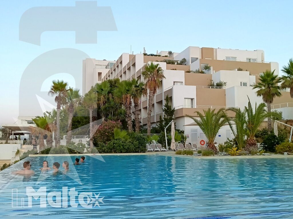Studenti in acqua nella piscina panoramica, con palme, lettini e struttura ricettiva moderna sullo sfondo.