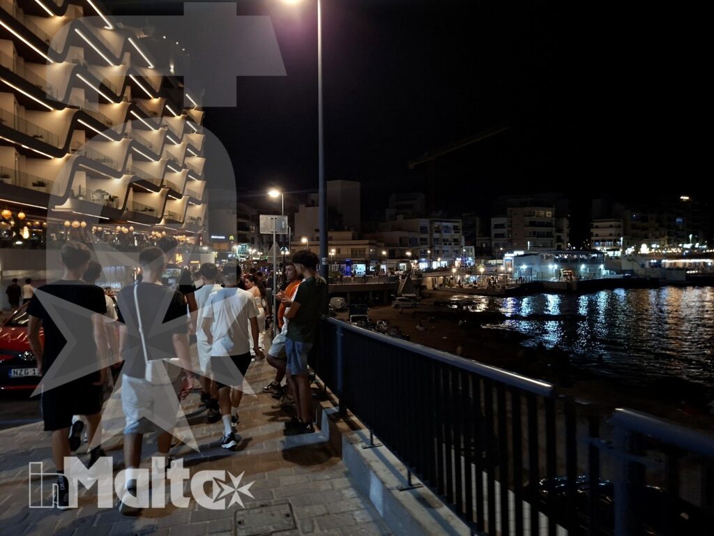 Un gruppo di studenti percorre il porto illuminato, tra riflessi sull'acqua, edifici e locali del waterfront maltese.
