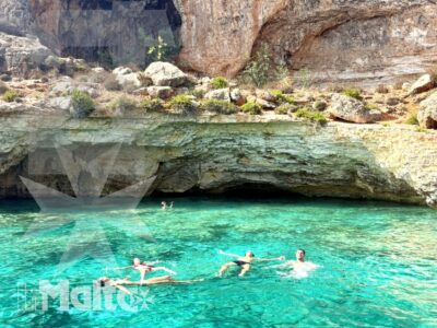 Gruppo di studenti nuota davanti a una parete rocciosa e a una grotta marina dalle acque turchesi, durante un'escursione a Malta.