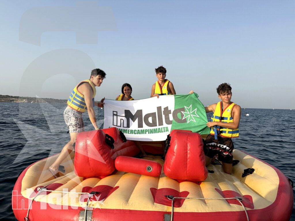 Quattro giovani con giubbotti salvagente posano su un gommone sostenendo uno striscione, con mare aperto e costa sullo sfondo.