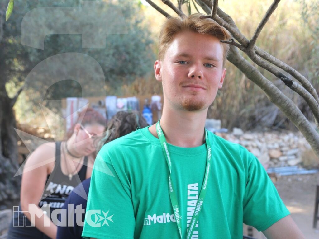 Studente con tesserino identificativo ritratto sotto un ulivo, mentre altri partecipanti svolgono attività all'aperto a Malta.