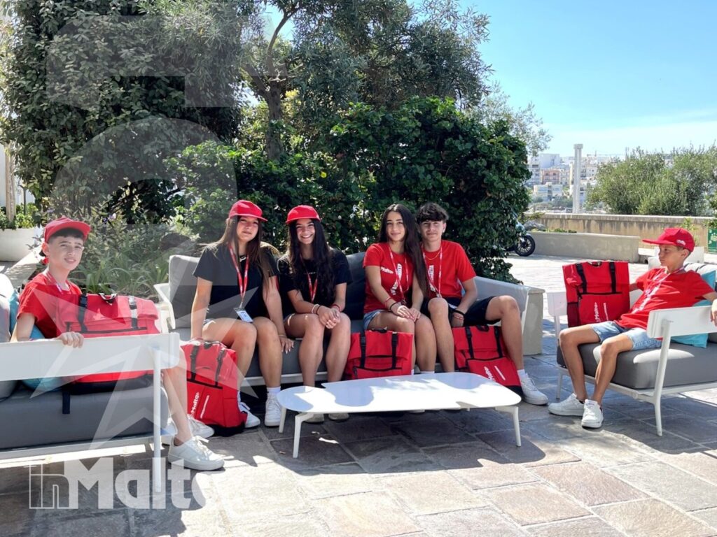 Studenti internazionali si rilassano tra divani e poltrone rossi in un patio esterno, con valigie e vegetazione mediterranea sullo sfondo.