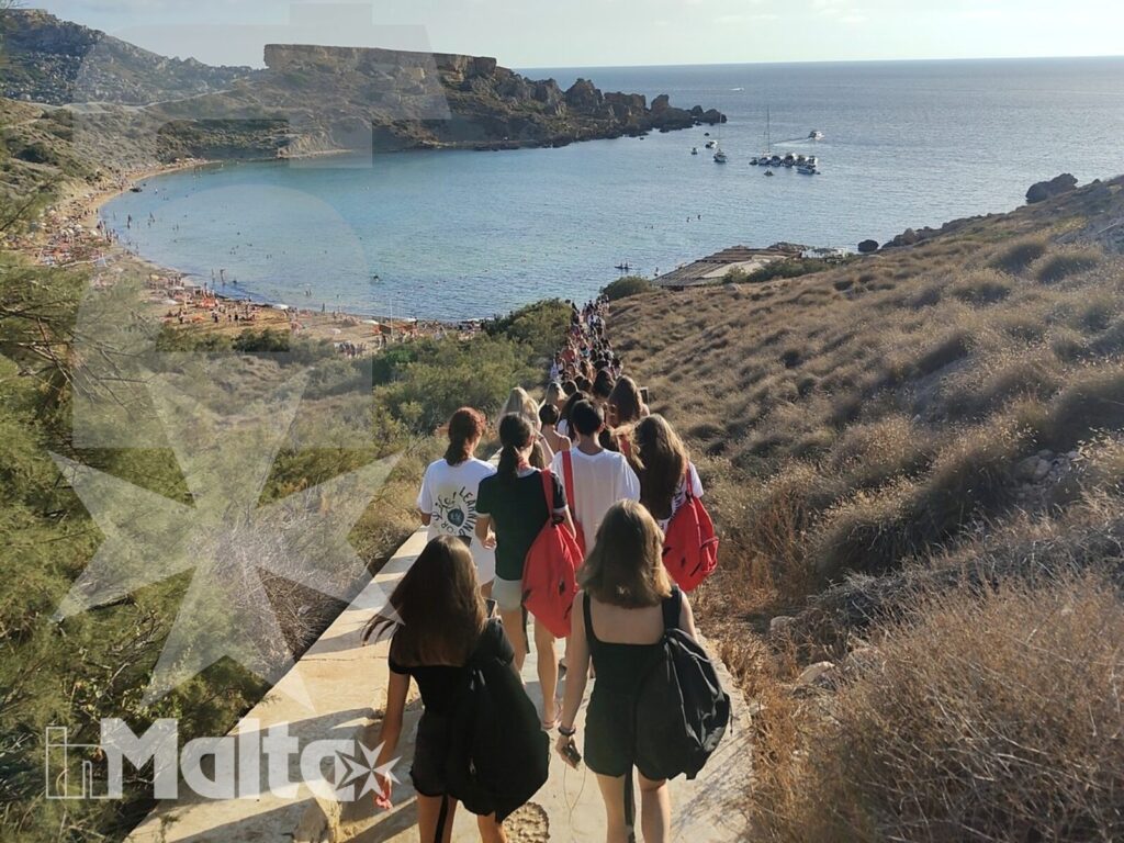 Studenti in escursione verso una baia di Malta Studenti percorrono un sentiero costiero panoramico verso una baia dall'acqua turchese, tra rocce e paesaggio mediterraneo.